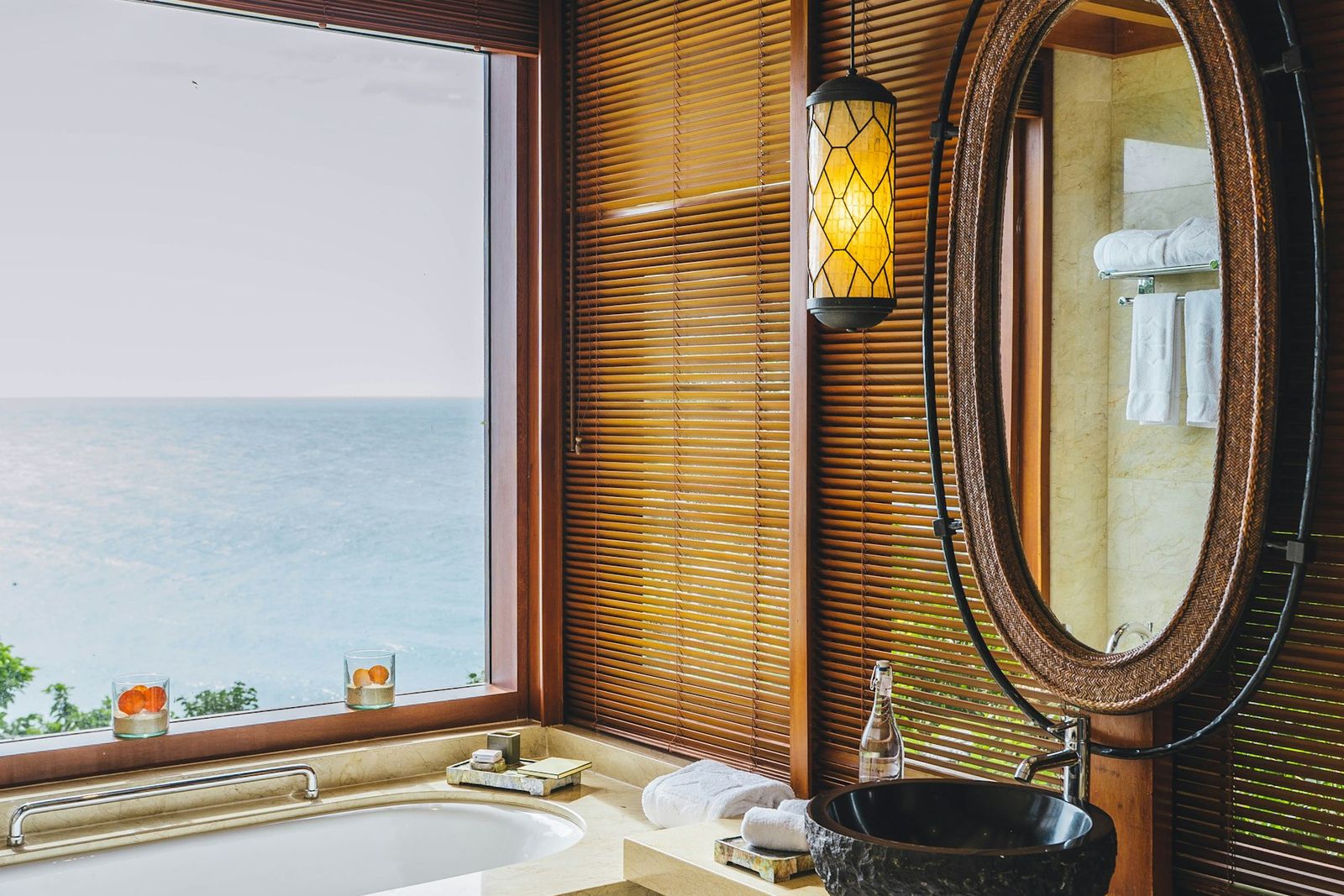 Bath beneath a wide window over the sea, with wooden blinds and an oval mirror