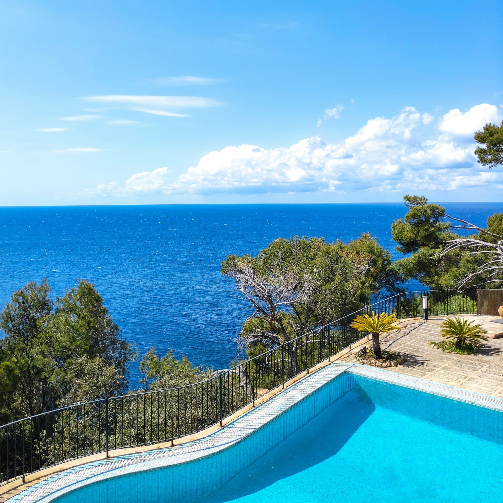 Blue pool on a stone terrace with pine trees and a wide view of the sea